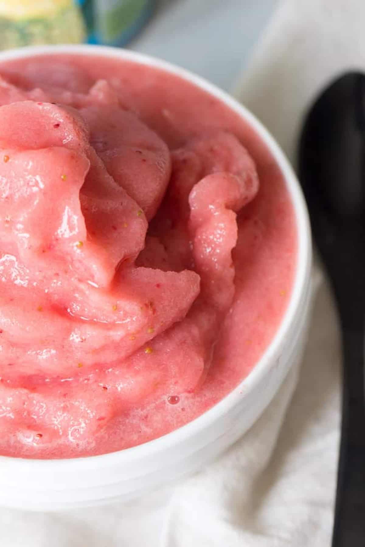 A white bowl filled with pink strawberry sorbet accompanied by a black spoon resting nearby on a white surface.