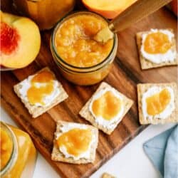 Jars of peach jam with a knife, surrounded by crackers topped with cream cheese and peach jam on a wooden board.
