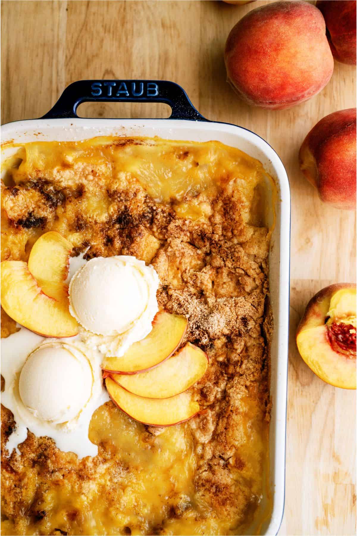 A baked peach cobbler in a Staub dish, topped with two scoops of vanilla ice cream and fresh peach slices, with whole peaches nearby on a wooden surface.