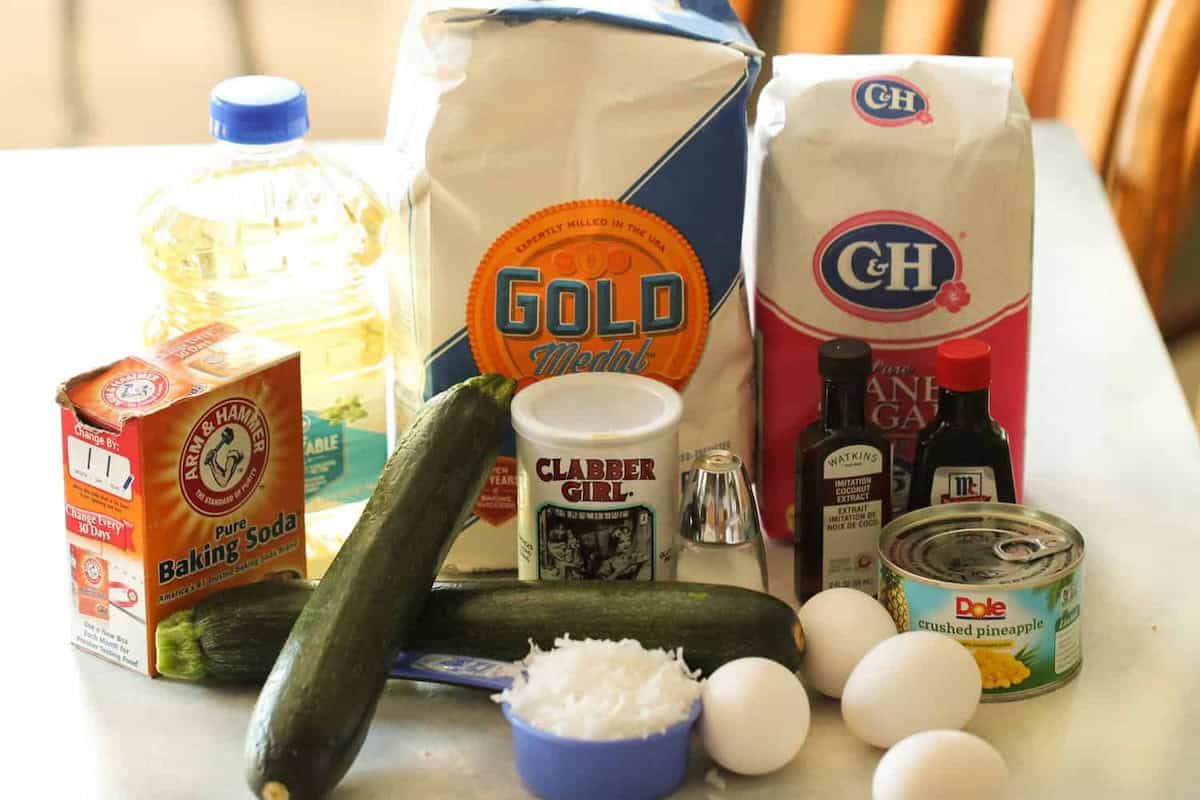 A variety of baking ingredients including flour, sugar, oil, baking soda, baking powder, vanilla extract, imitation rum flavor, eggs, crushed pineapple, zucchini, and shredded coconut on a table.