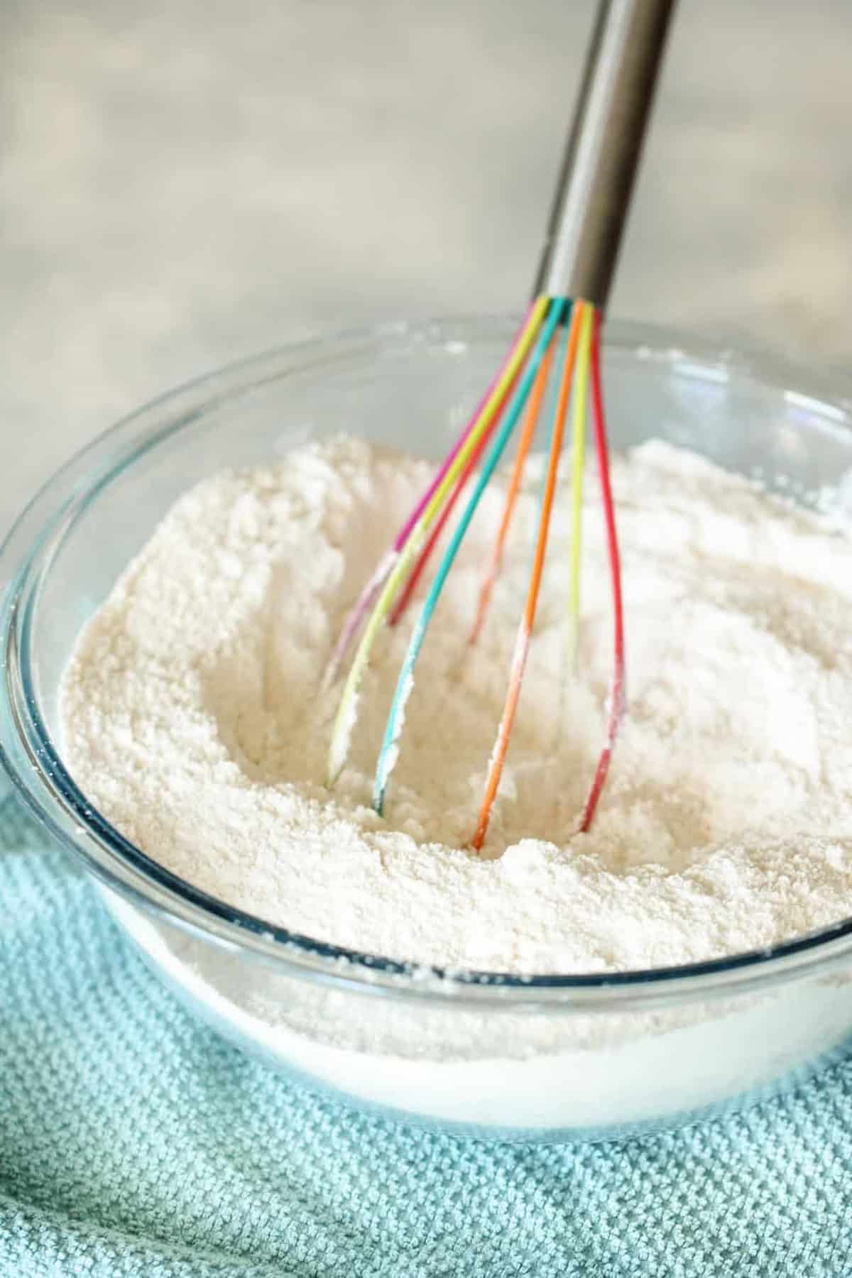 A whisk with a multi-colored handle is in a glass mixing bowl containing flour, placed on a light blue textured cloth.