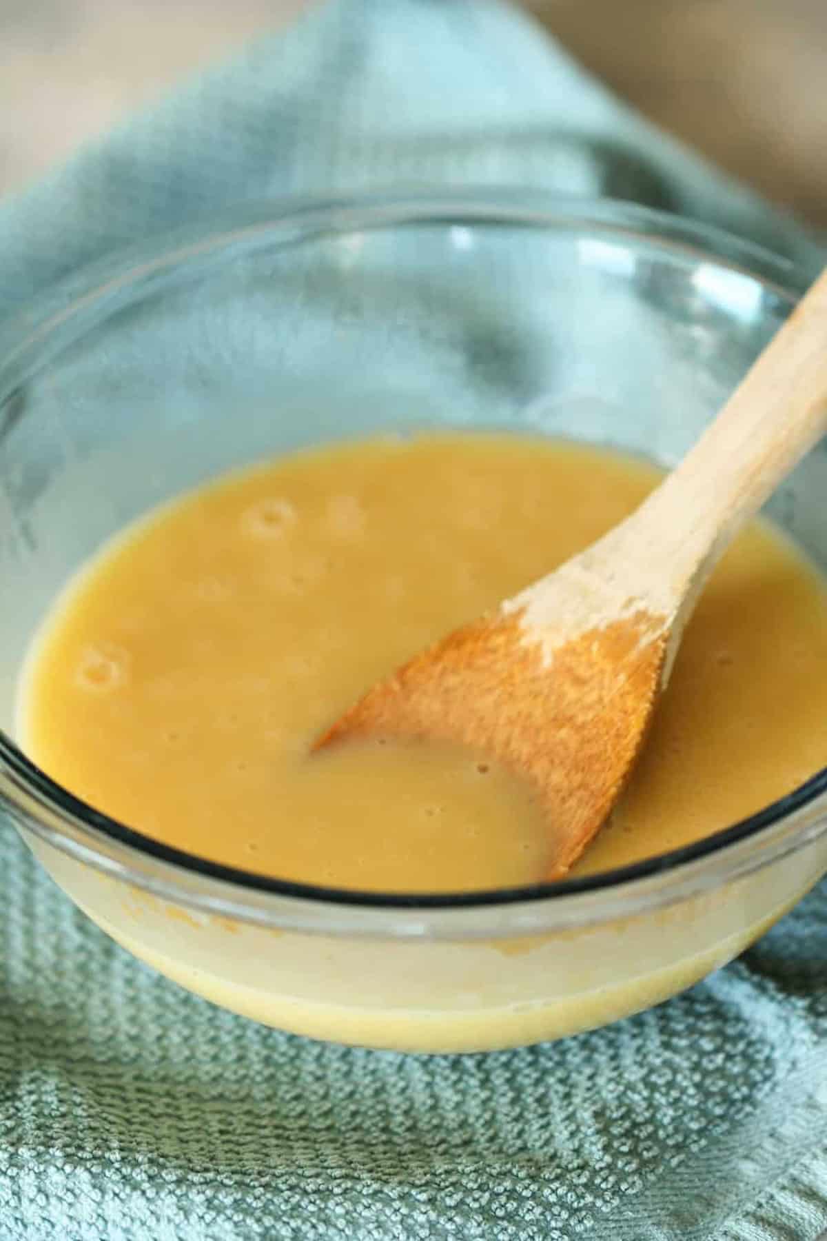 A glass bowl containing beige liquid batter, with a wooden spoon partially submerged, resting on a light blue textured cloth.