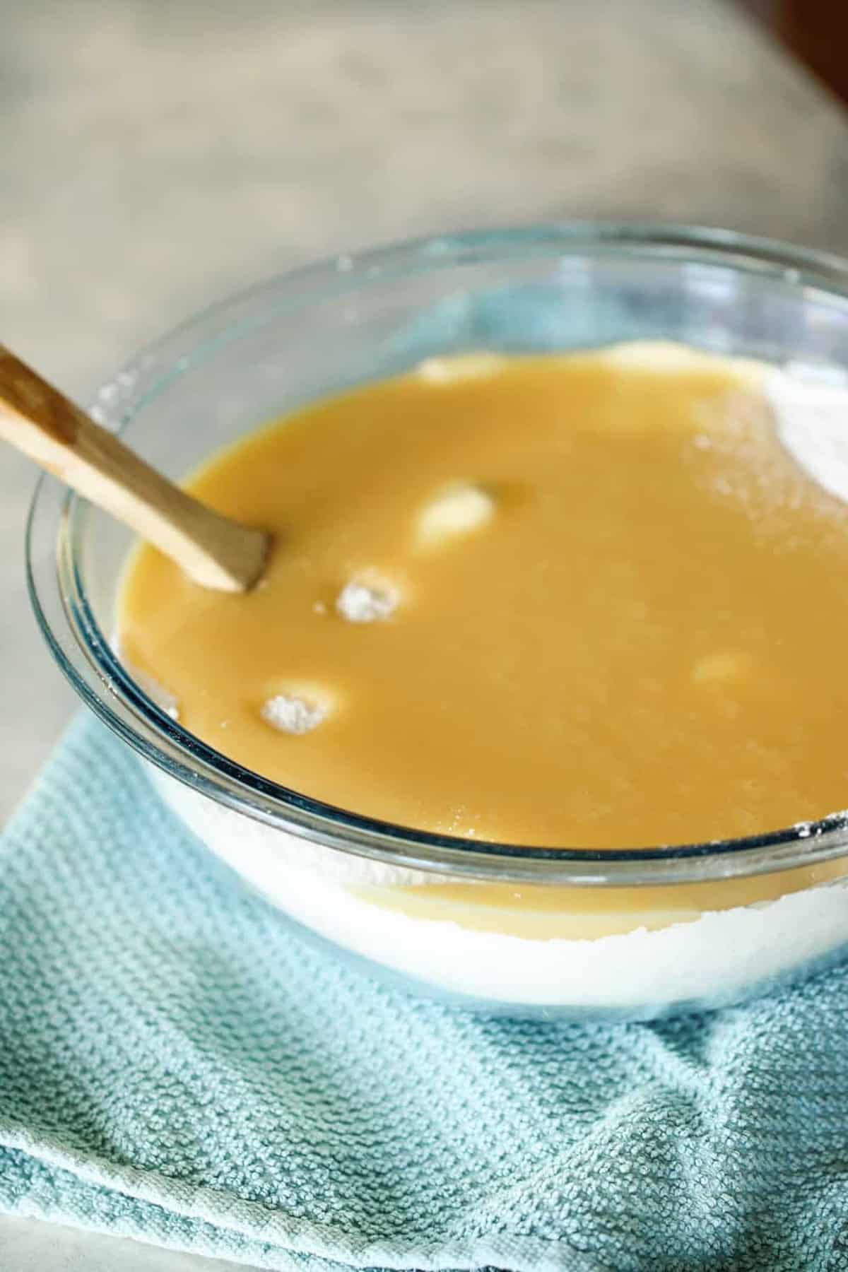 A glass bowl filled with a creamy yellow batter and a wooden spoon in it sits on a light blue cloth.