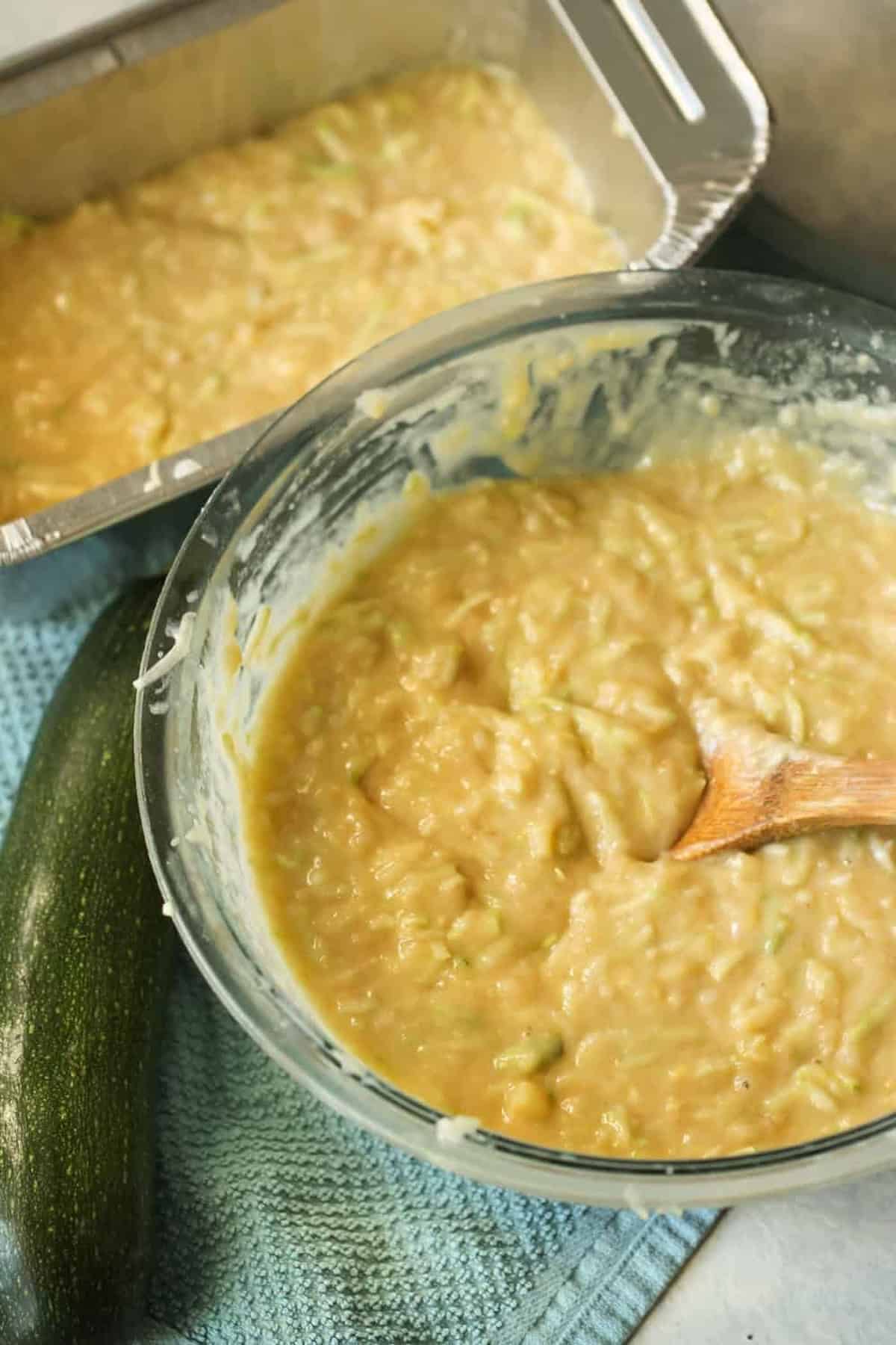 A bowl of pale yellow batter being mixed with a wooden spoon sits beside a zucchini and a tray of the same batter on a blue towel.