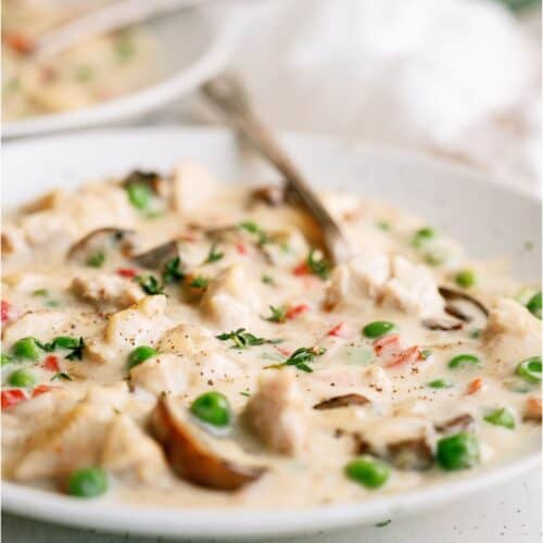 A creamy chicken and vegetable stew in a white bowl with peas, mushrooms, and red bell peppers, garnished with herbs, with a spoon in the dish.