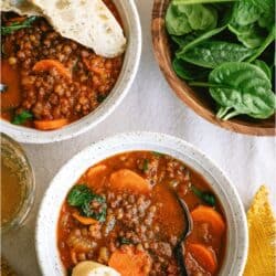Two bowls of lentil and vegetable soup with carrot slices and bread, next to a wooden bowl of fresh spinach leaves and two glasses of drink.