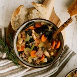 A bowl of soup with ground meat, white beans, carrots, celery, and greens sits on a striped towel next to slices of bread and a wooden cutting board.