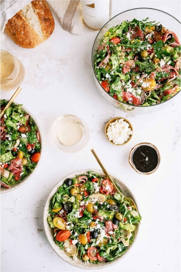Top view of 3 bowls of Antipasto Salad with a loaf of bread and glasses surrounding the bowls.