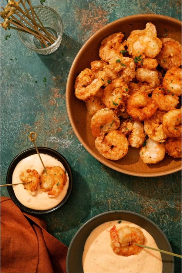 A bowl of cooked shrimp with herbs, beside two small bowls of dipping sauce and skewered shrimp on a green textured surface.