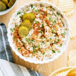 A white bowl of Fried Pickle Dip surrounded by potato chips and a small bowl of pickles in the background.