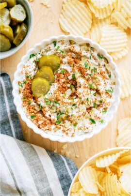 A white bowl of Fried Pickle Dip surrounded by potato chips and a small bowl of pickles in the background.