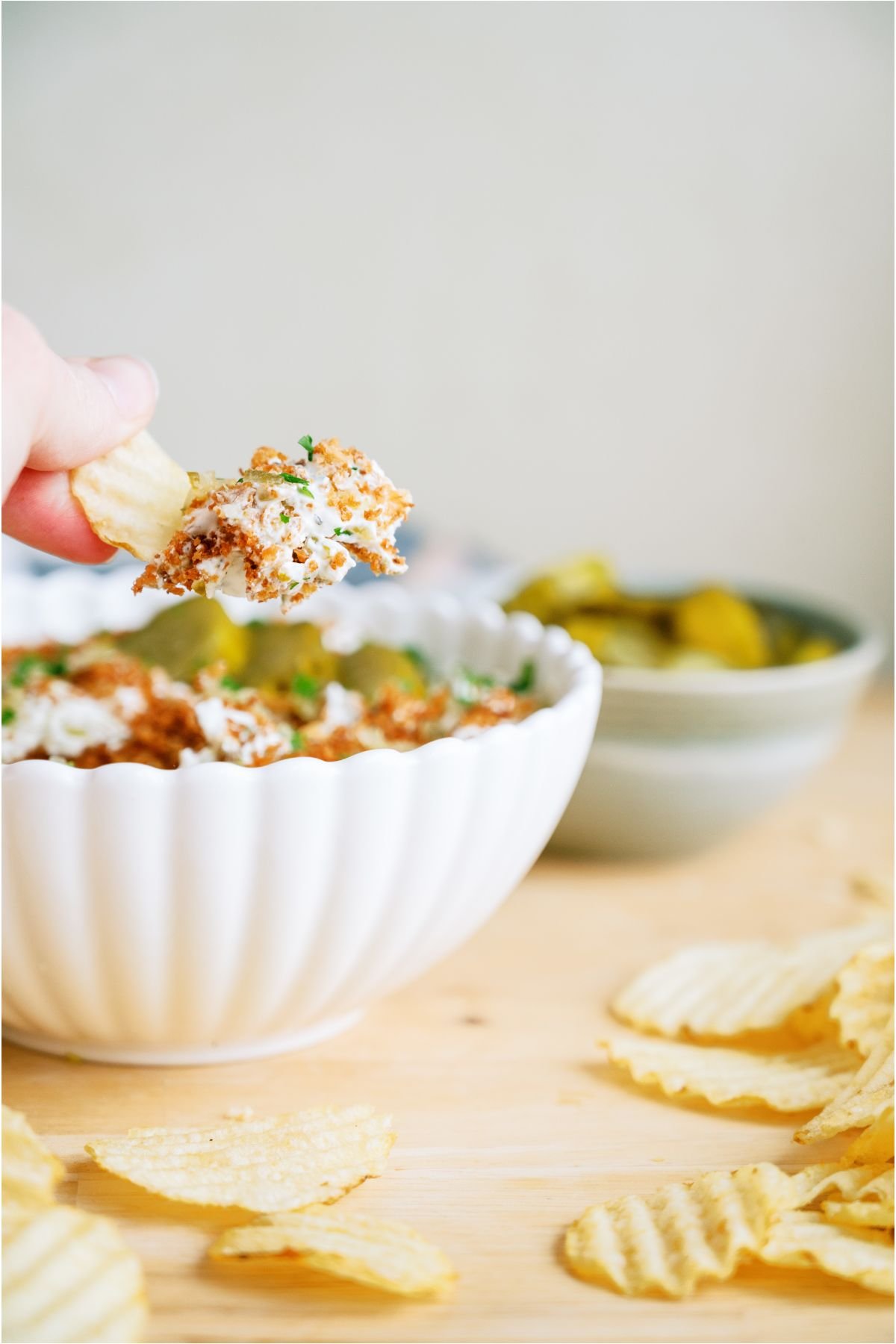 A hand lifting a chip with Fried Pickle Dip on it out of a bowl of Fried Pickle Dip with potato chips on the side.