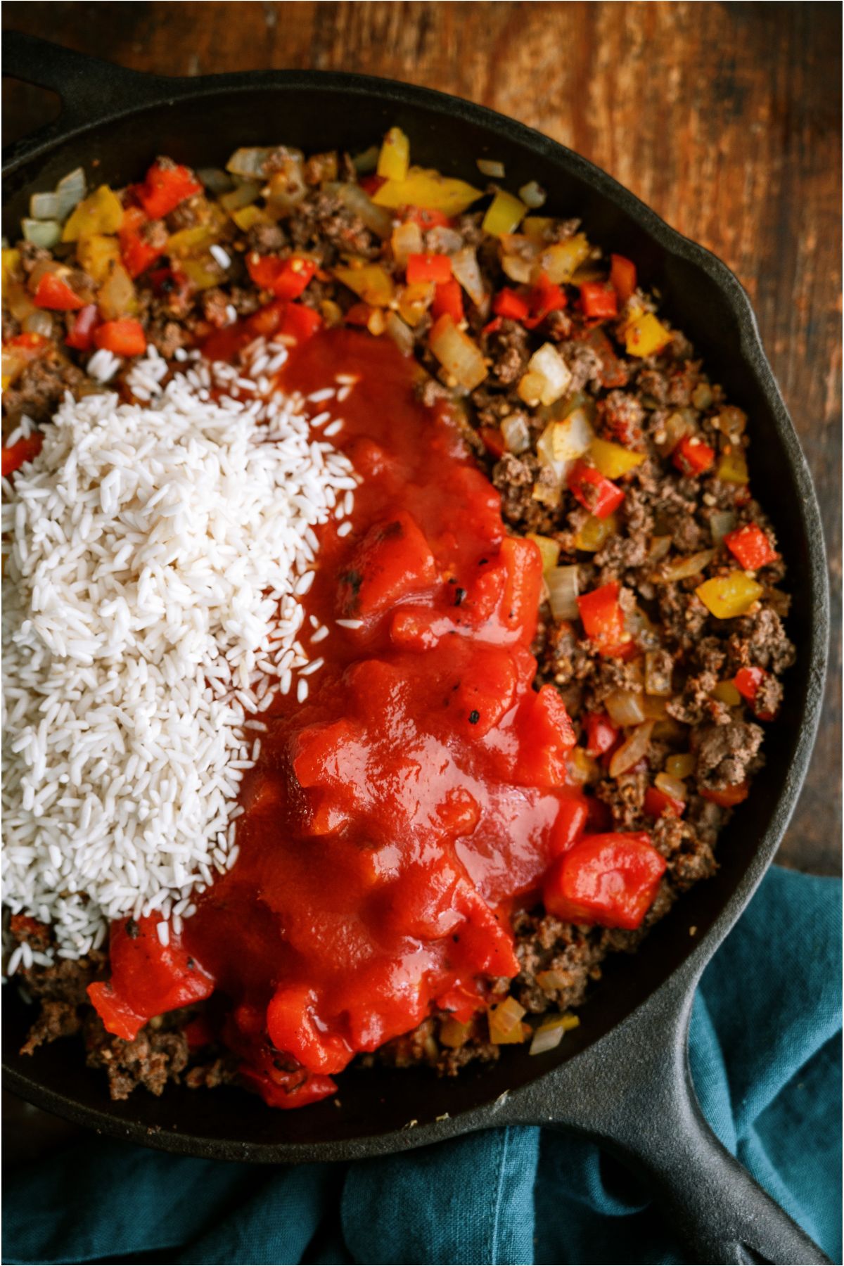 A skillet with ground beef, onions and peppers with diced tomatoes and rice on top.