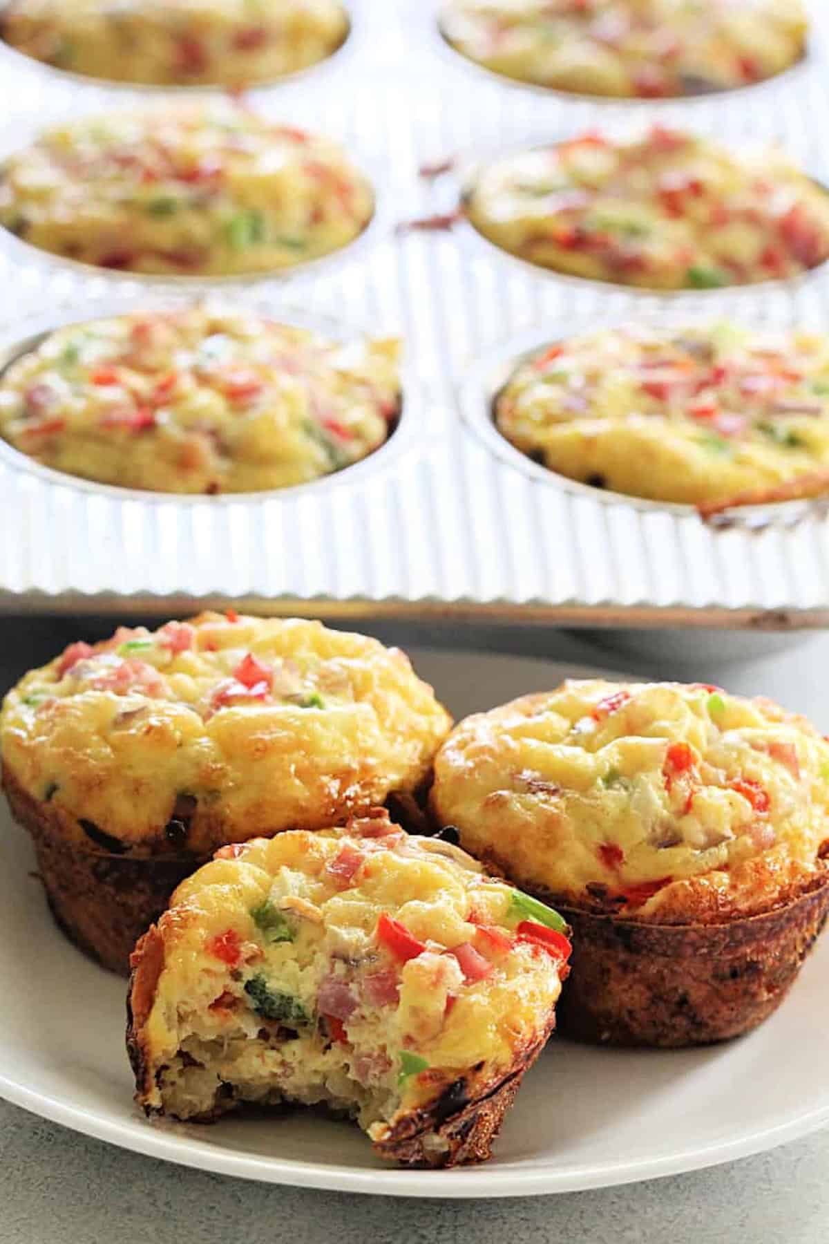 A plate with three egg muffins, one partially eaten, filled with vegetables and bits of meat. A muffin tray with more egg muffins sits in the background.