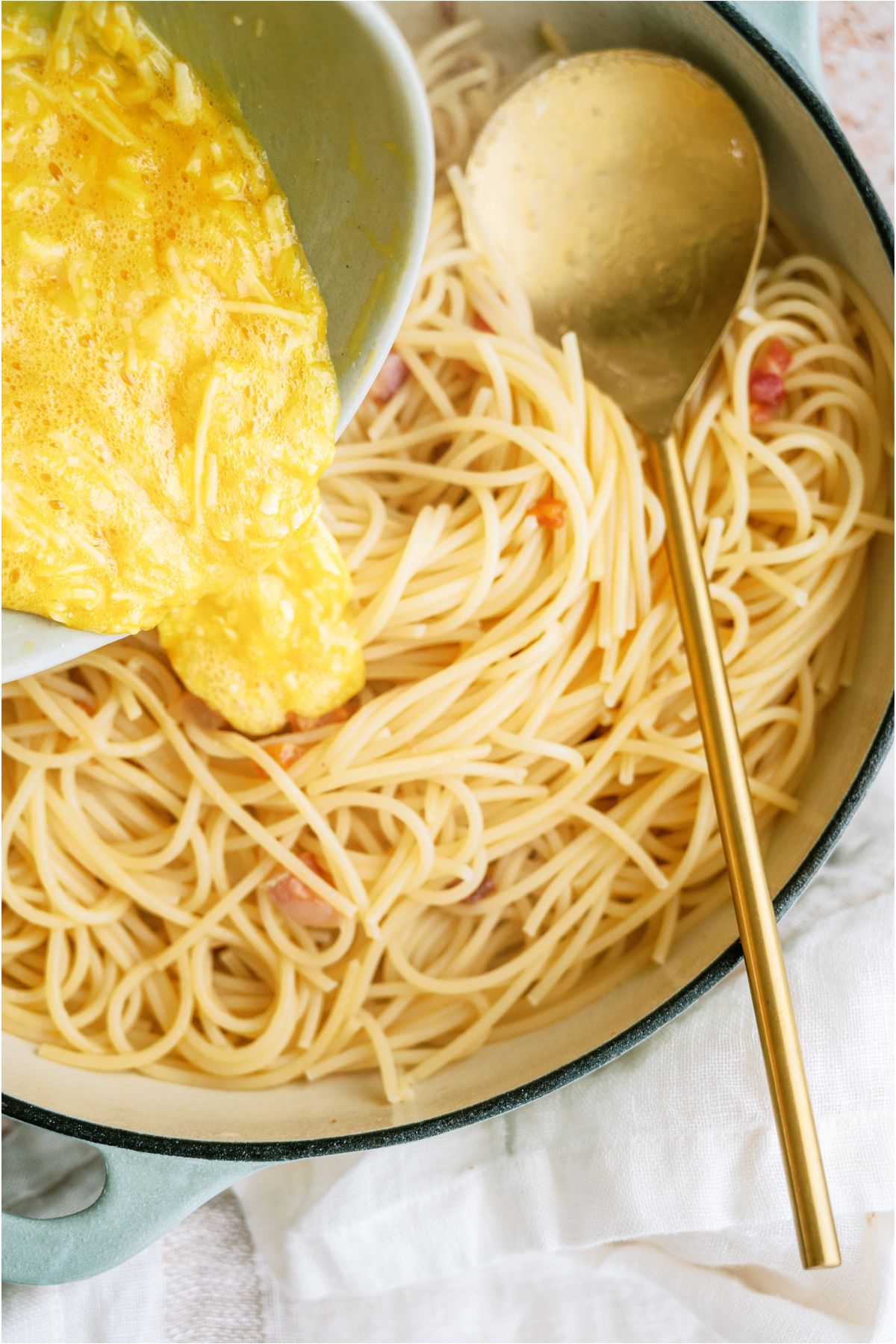 Pouring egg and parmesan mixture into a pan of cooked spaghetti noodles.