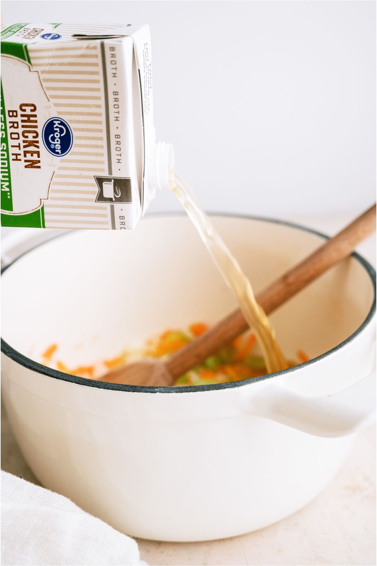 Pouring chicken broth from a carton into a large stockpot with chopped vegetables and a wooden spoon.
