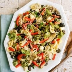 A platter of salad with tortellini, artichokes, bell peppers, olives, herbs, and grated cheese on a tiled surface with a blue napkin and wooden utensils beside it.