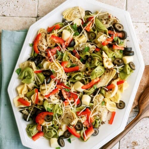 A platter of salad with tortellini, artichokes, bell peppers, olives, herbs, and grated cheese on a tiled surface with a blue napkin and wooden utensils beside it.