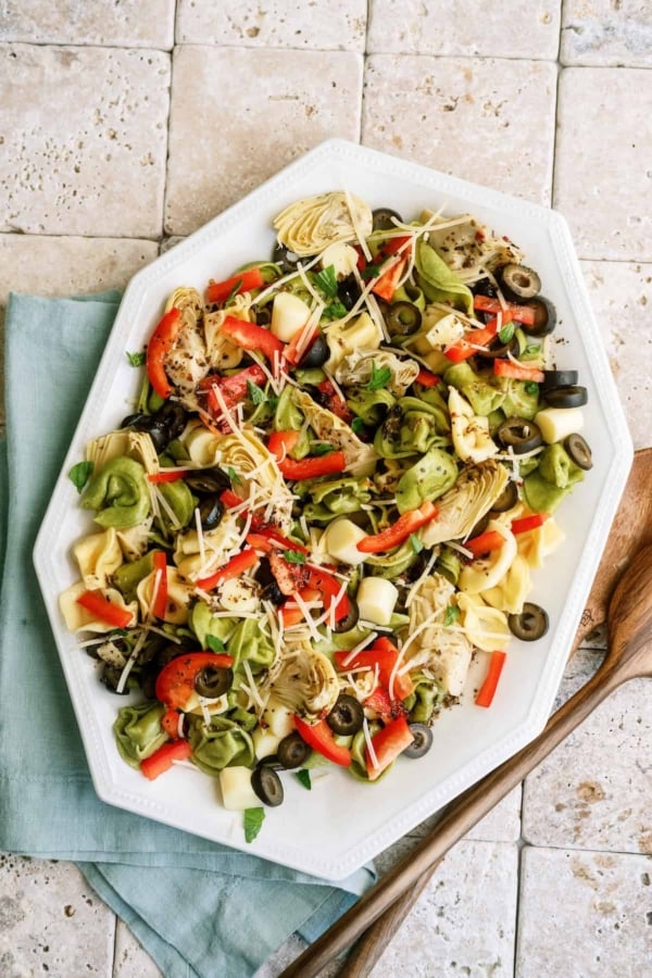 A platter of salad with tortellini, artichokes, bell peppers, olives, herbs, and grated cheese on a tiled surface with a blue napkin and wooden utensils beside it.