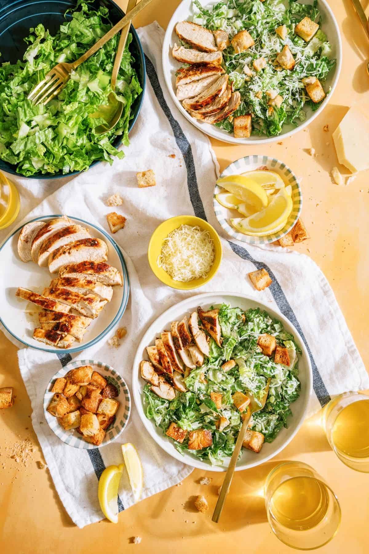 Top view of 2 Grilled Chicken Caesar Salads in bowls, with a bowl of salad, a plate of grilled chicken, small bowl of lemon wedges and a small bowl of shredded cheese.