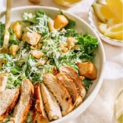 A bowl with Caesar salad topped with grilled chicken. A small bowl of shredded cheese in the background.