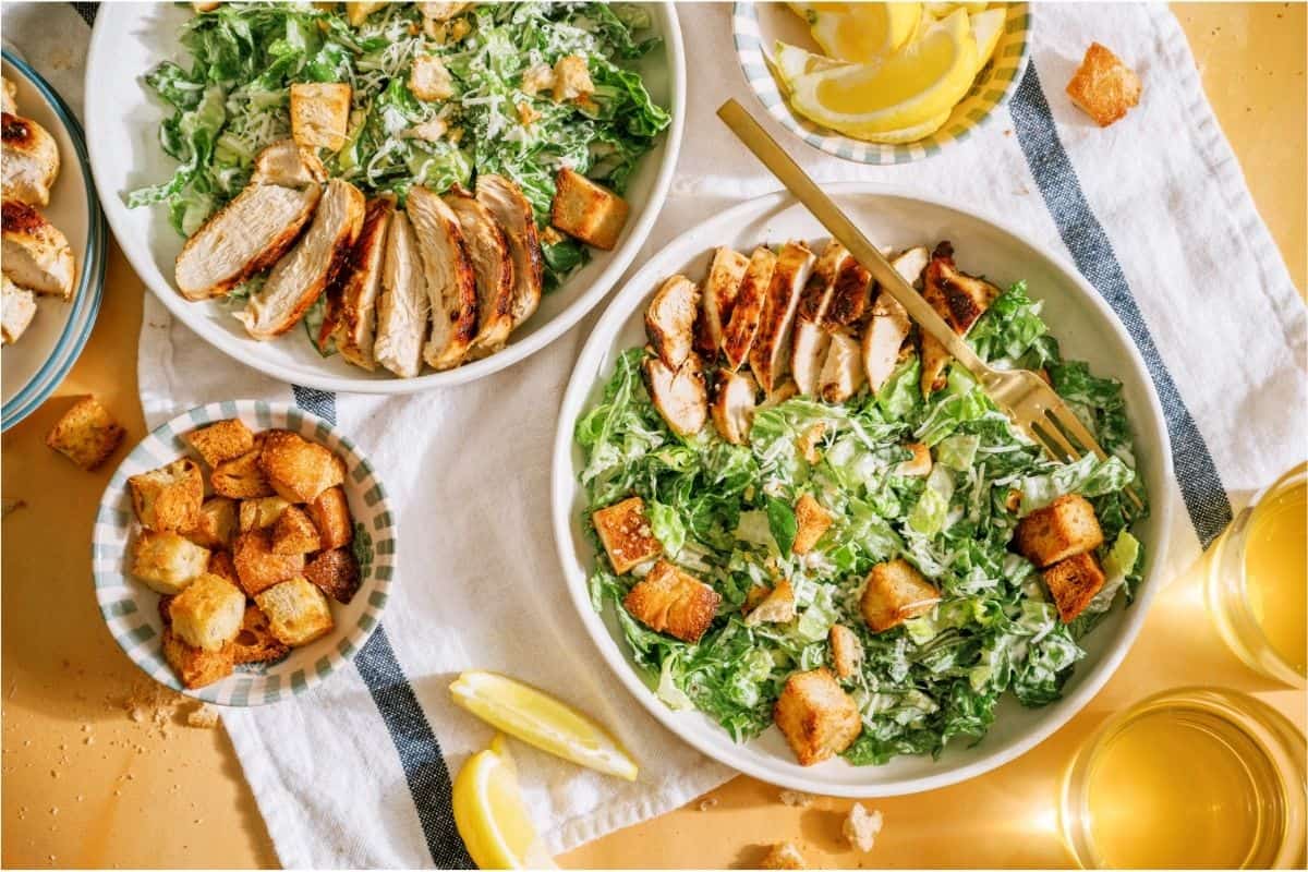 2 bowls of Grilled Chicken Caesar Salad with a small bowls of croutons and lemon wedges off to the side.