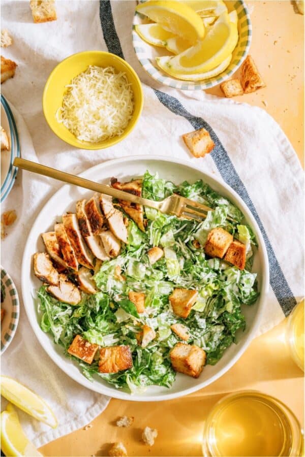 A bowl of Caesar salad with grilled chicken, croutons, and dressing, surrounded by shredded cheese, lemon wedges, and glasses of yellow beverage on a table.