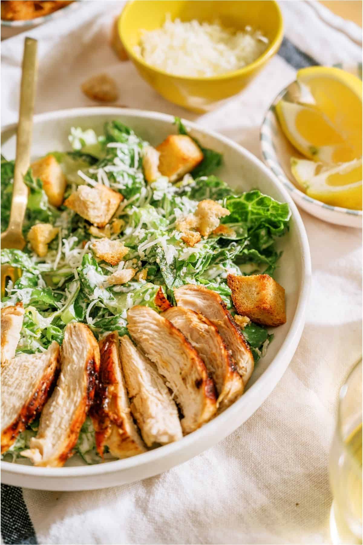 A bowl with Caesar salad topped with grilled chicken. A small bowl of shredded cheese in the background.
