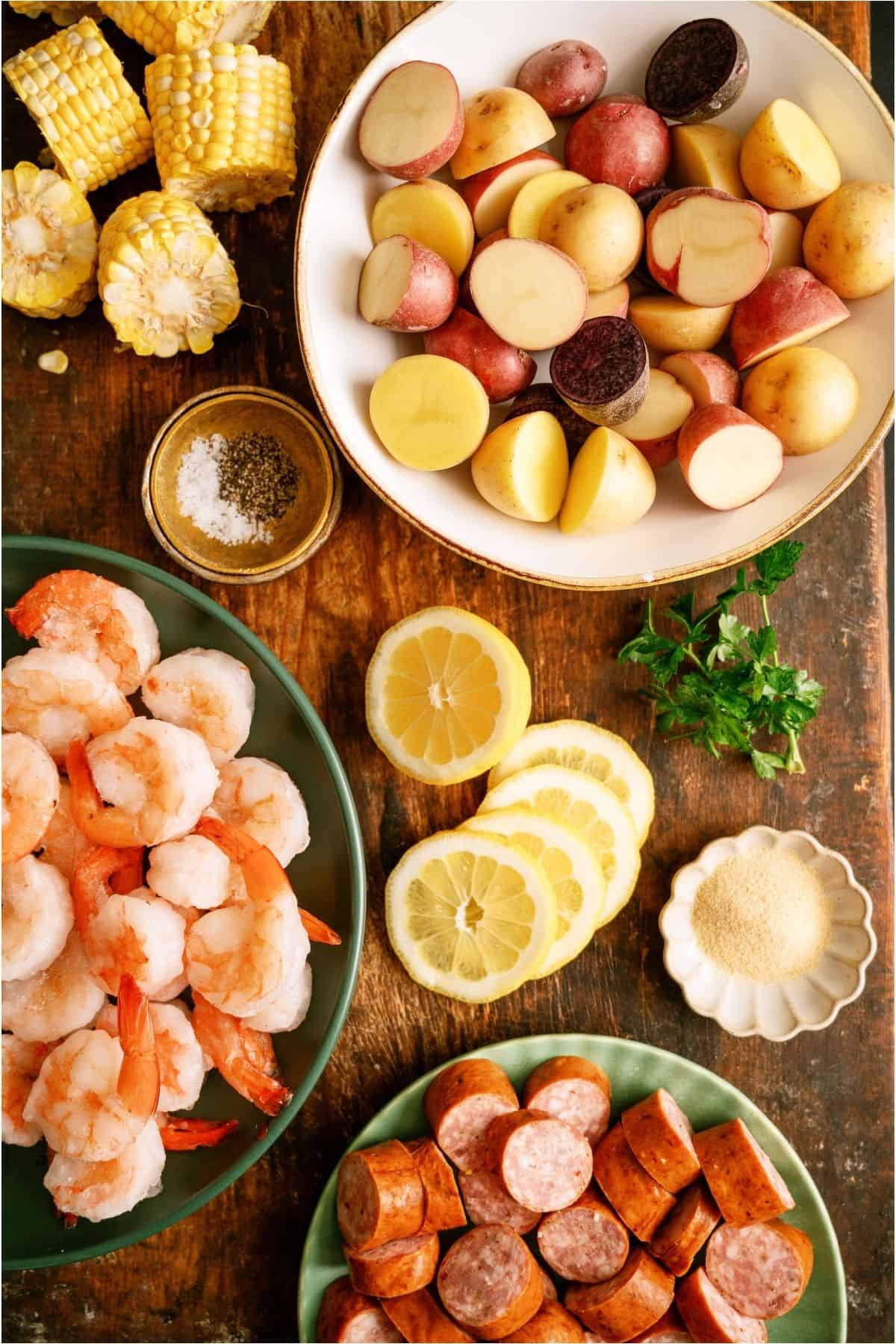 A top-down view of raw ingredients on a wooden table, including shrimp, sausage, sliced potatoes, corn, lemon, parsley, garlic powder, and a bowl of salt and pepper.