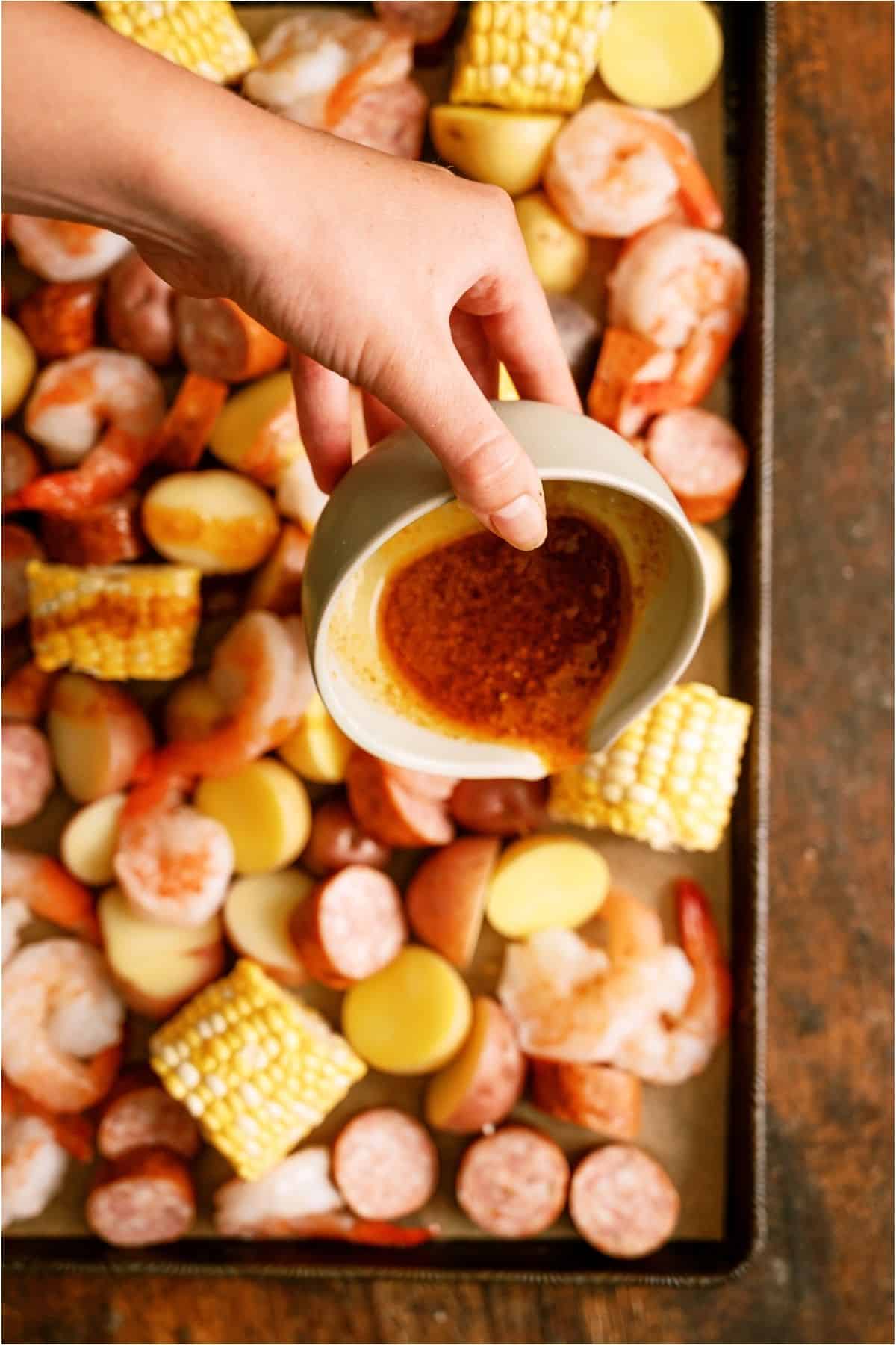 A hand pours seasoned butter from a small bowl onto a sheet pan filled with shrimp, sausage, potato slices, and corn pieces.