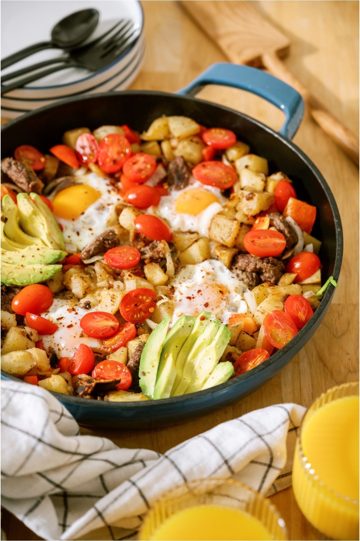 A skillet of Steak And Eggs with veggies, topped with avocado. A stack of plates and silverware in the background.