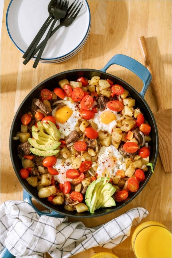 A pan of Steak And Eggs with veggies, topped with avocado. A stack of plates and silverware in the background.
