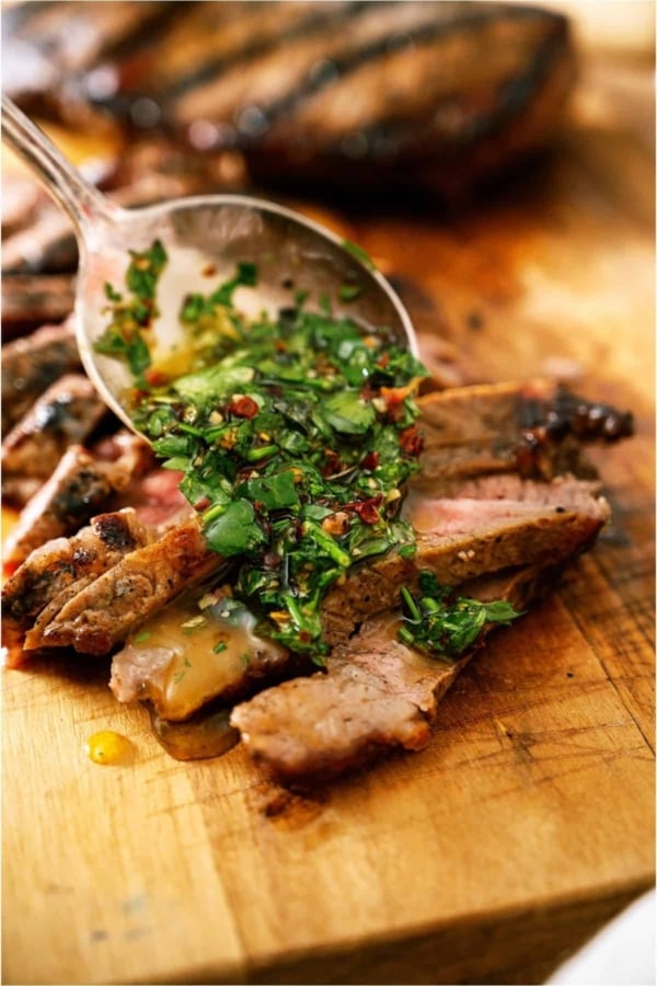 Chimichurri Sauce being spooned over sliced steak on a cutting board.