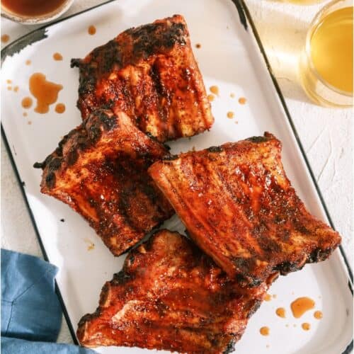 Four grilled pork rib racks with barbecue sauce on a white tray, with two glasses of light-colored beverage and a blue napkin nearby.