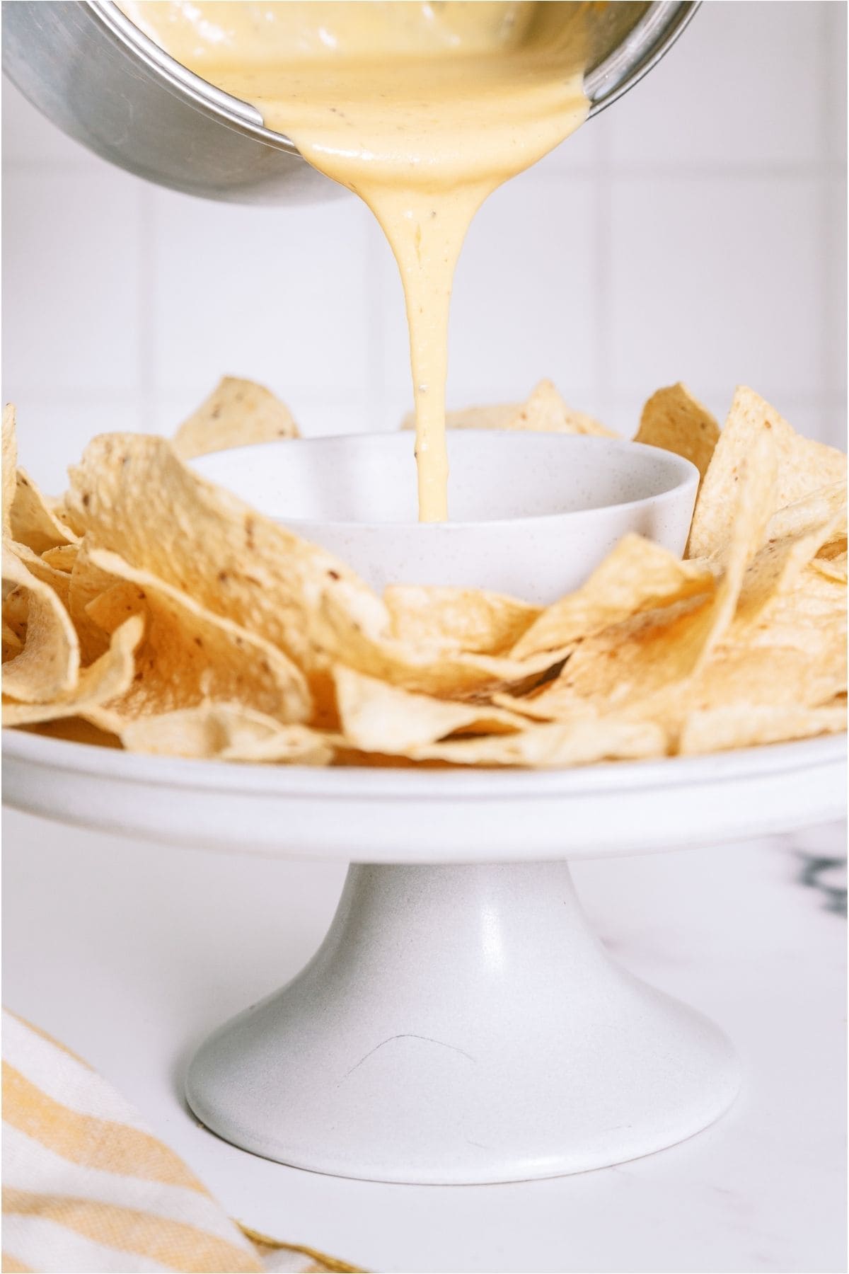 Pouring Quick Nacho Cheese Sauce into a bowl surrounded by tortilla chips.