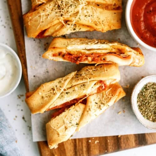 Sliced braided bread filled with cheese and tomato sauce, topped with herbs, served on parchment paper with bowls of marinara, ranch, and dried herbs.