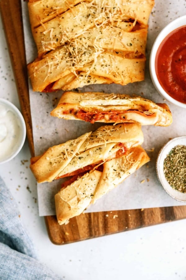 Sliced braided bread filled with cheese and tomato sauce, topped with herbs, served on parchment paper with bowls of marinara, ranch, and dried herbs.