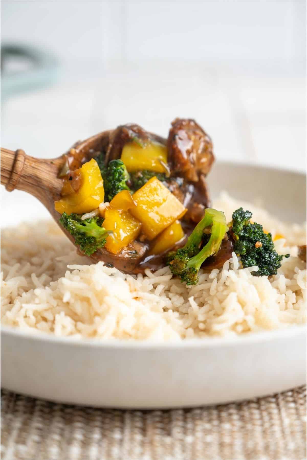 A wooden spoon placing a scoop of Chicken And Sausage Stir Fry over rice on a plate.