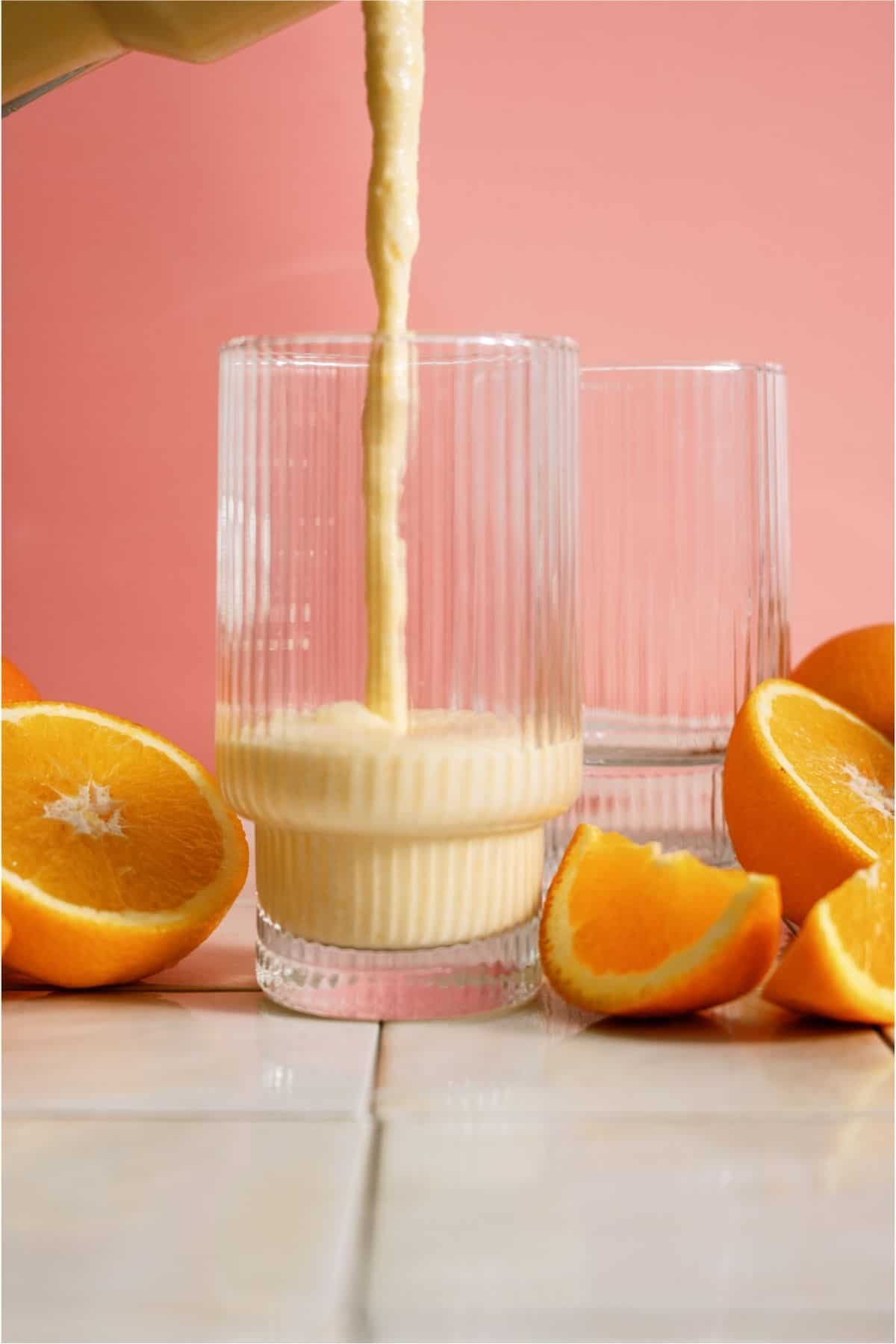A blender pouring Copycat Orange Julius in a cup surrounded by oranges.
