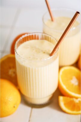 Orange julius smoothie in a cup surrounded with a straw and oranges.