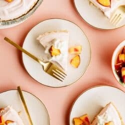 Five slices of peach pie topped with whipped cream and peach chunks are served on plates with gold forks, arranged on a peach-colored surface.