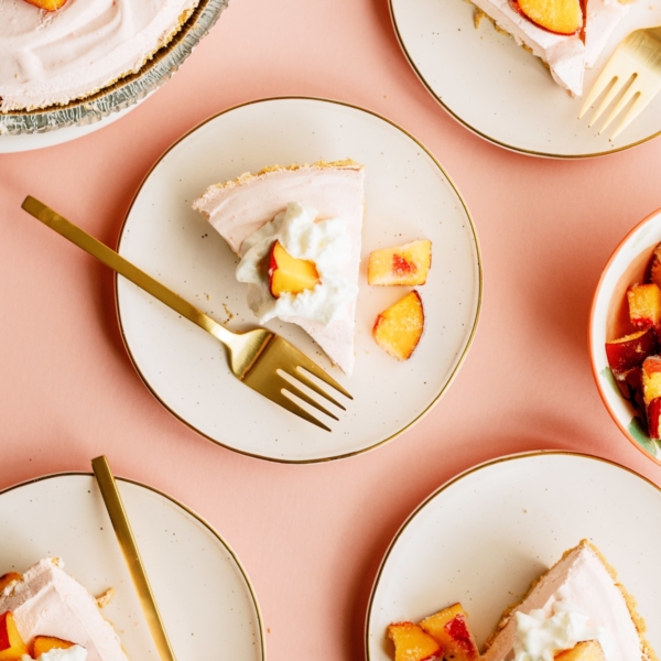 Five slices of peach pie topped with whipped cream and peach chunks are served on plates with gold forks, arranged on a peach-colored surface.