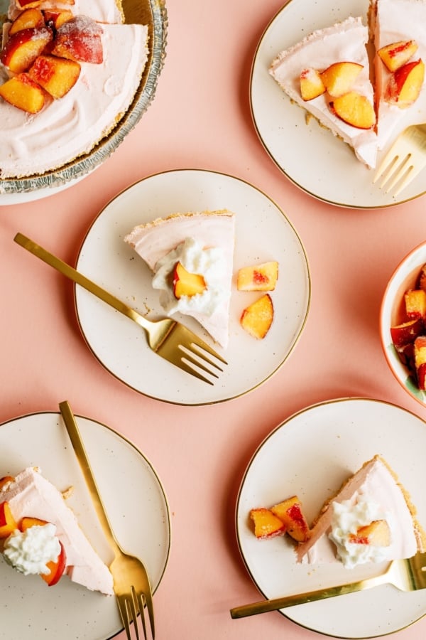 Five slices of peach pie topped with whipped cream and peach chunks are served on plates with gold forks, arranged on a peach-colored surface.
