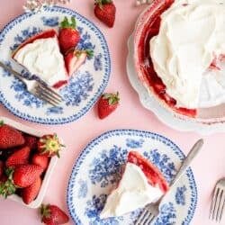 Overhead view of two plates with slices of strawberry pie topped with whipped cream, a whole pie, fresh strawberries, and white flowers on a pink surface.