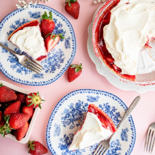 Overhead view of two plates with slices of strawberry pie topped with whipped cream, a whole pie, fresh strawberries, and white flowers on a pink surface.