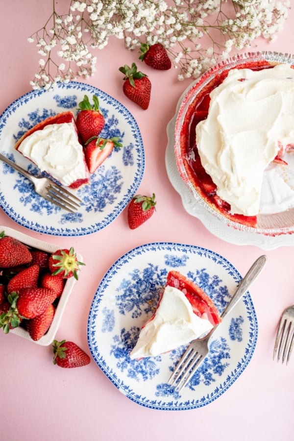 Overhead view of two plates with slices of strawberry pie topped with whipped cream, a whole pie, fresh strawberries, and white flowers on a pink surface.
