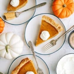 Three plates each with a slice of pumpkin pie topped with whipped cream, surrounded by small pumpkins and a bowl of whipped topping.