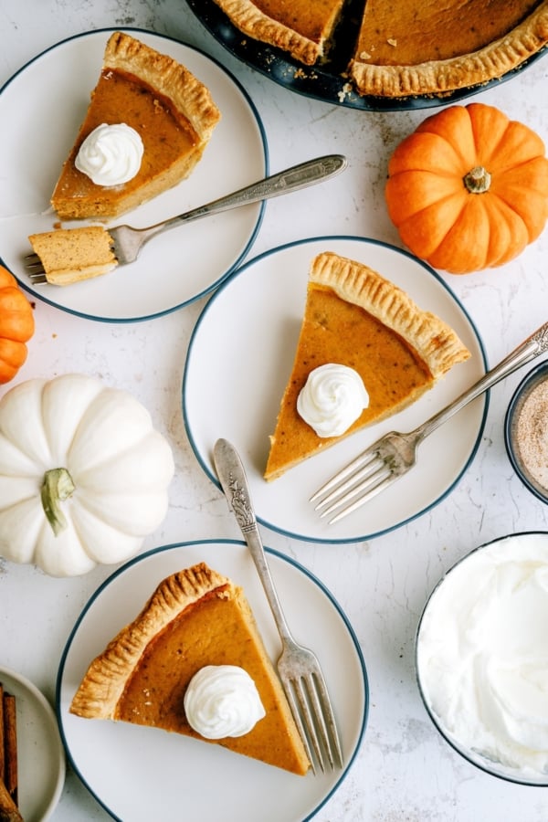 Three plates each with a slice of pumpkin pie topped with whipped cream, surrounded by small pumpkins and a bowl of whipped topping.