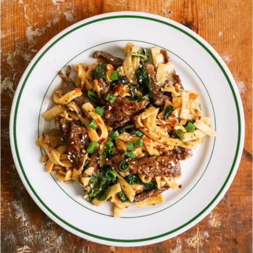A servings of Beef and Noodles in a white bowl on a wooden background.