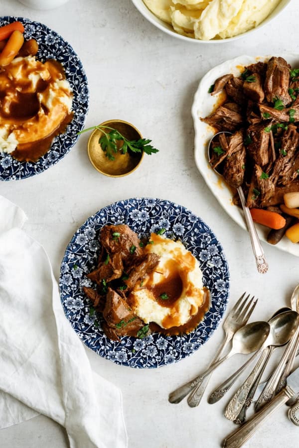 Two blue-patterned plates with pot roast, carrots, and mashed potatoes with gravy, alongside a serving platter of pot roast, a bowl of mashed potatoes, and assorted silverware on a white table.